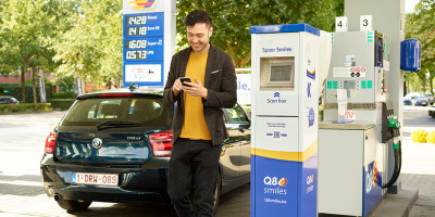 Un homme s'apprêtant à encaisser ses points/Smiles tout en regardant son portable à côté de sa voiture bleue à la pompe.